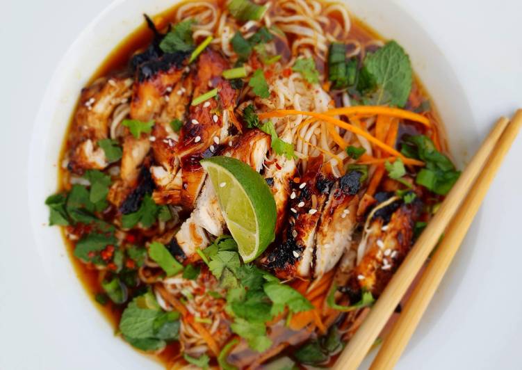 Overhead photograph of a noodle soup bowl with grilled meat, lime wedge, herbs and shredded carrot, with chopsticks resting on a wooden surface, warm amber and green tones.
